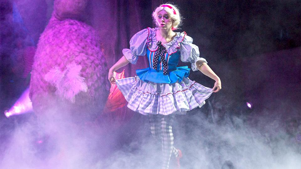 An actress portraying Alice walks through fog on stage while holding her skirt out